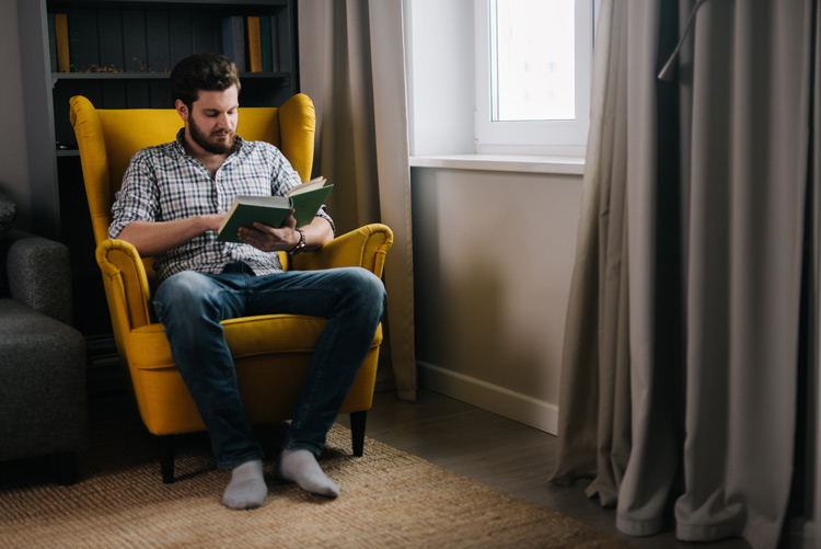 man sitting at home in yellow chair reading a book - mental health - COVID-19