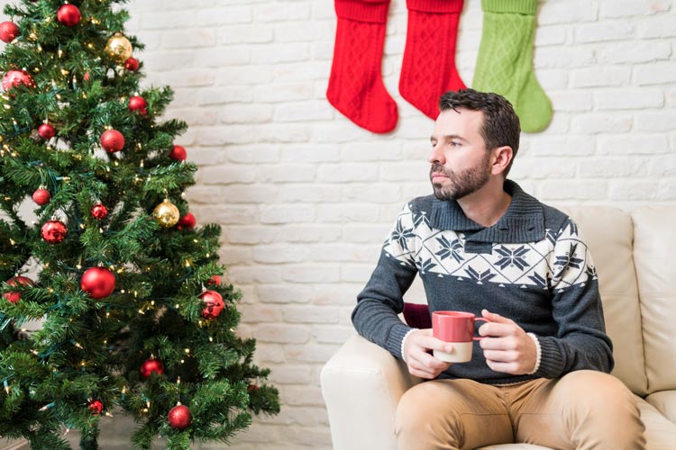 man in his thirties sitting alone next to Christmas tree drinking coffee or cocoa, thinking - loneliness
