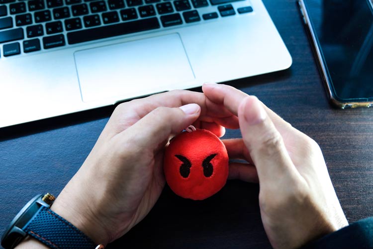 closely cropped shot of hands holding a red stress toy with a frowning face on it - person is sitting at their desk in front of a laptop - anger management
